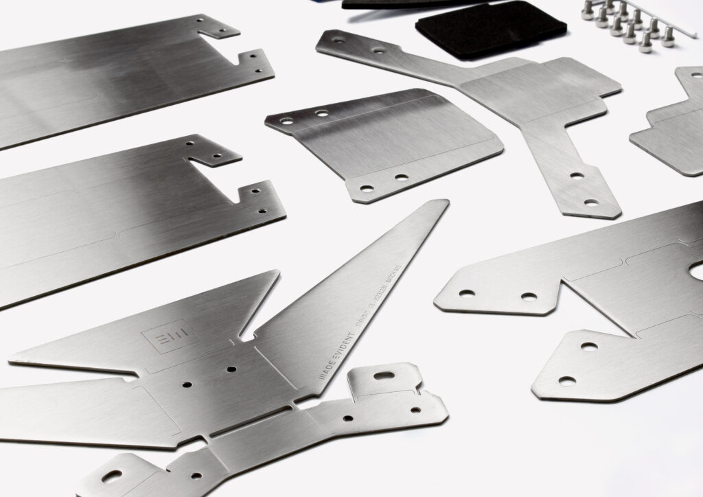 StandV1 - Best Imagery_0001_Layer 5 Several brushed metal plates and components with various cut-outs and shapes, likely designed using CAD, are laid out on a white surface alongside screws and small black rectangular pads.