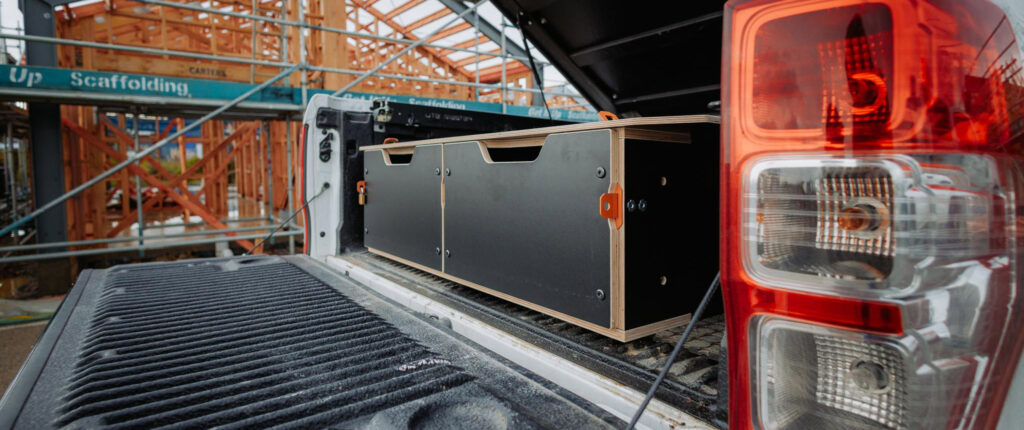 Workvan Ute Drawer A pickup truck with its tailgate down shows a black and wood storage drawer system, reflecting expert Product Design NZ. In the background, a building under construction with scaffolding is visible.