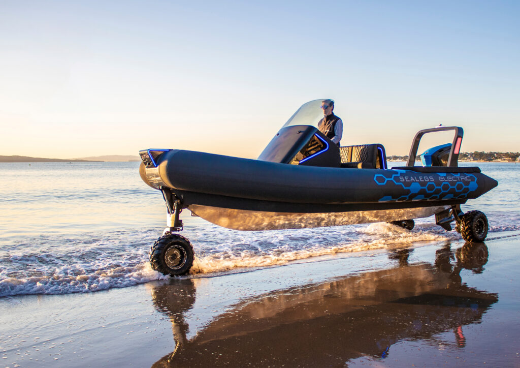 A person is driving an amphibious boat with wheels onto a sandy beach at sunset. The water is calm, the sky clear. Showcasing innovative Product Design NZ, the boat has SeaLegs Electric written on its side.