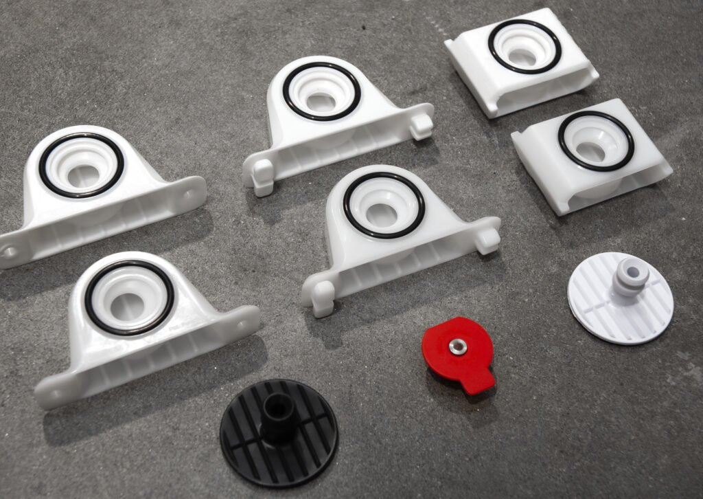 FAS009 - Best Award Stratlock HERO Several white and black plastic components with circular openings and rubber rings, plus one red and one white round part, are arranged on a grey surface—showcasing Industrial Design NZ expertise in CAD and design for manufacture.