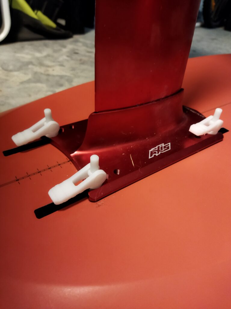 IMG_20220519_183646 Close-up of a red hydrofoil mast attached to a board with three white plastic fasteners, showcasing sleek Product Design NZ. The board surface is red with a small white logo near the base of the mast.