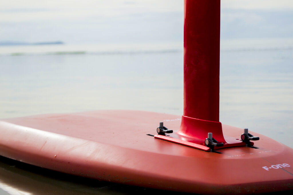 Close-up of a red hydrofoil surfboard resting on calm water, showing the base of the mast and smooth board surface with F-one written in white lettering. Product Design NZ expertise influences its sleek, CAD-enhanced lines. Out-of-focus shoreline and sky behind.