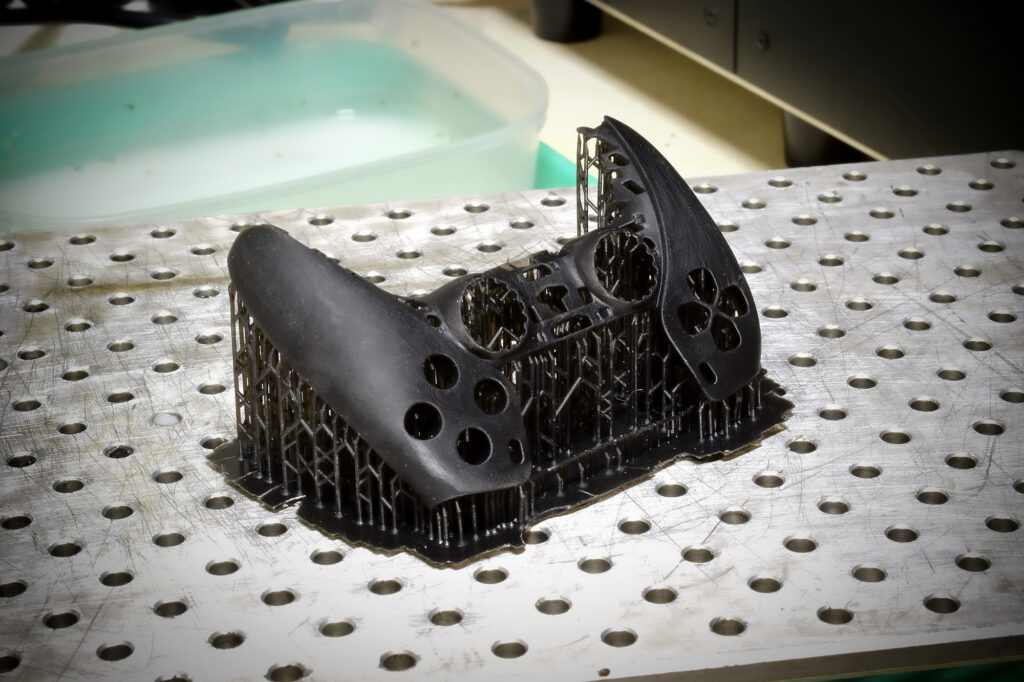 ET-ID Industrial Design 3D Print SLA FDM B A 3D-printed object resembling a game controller sits on a perforated metal surface, still attached to its support structures—a testament to thoughtful CAD and Design for Manufacture principles—with a tray of liquid in the background.