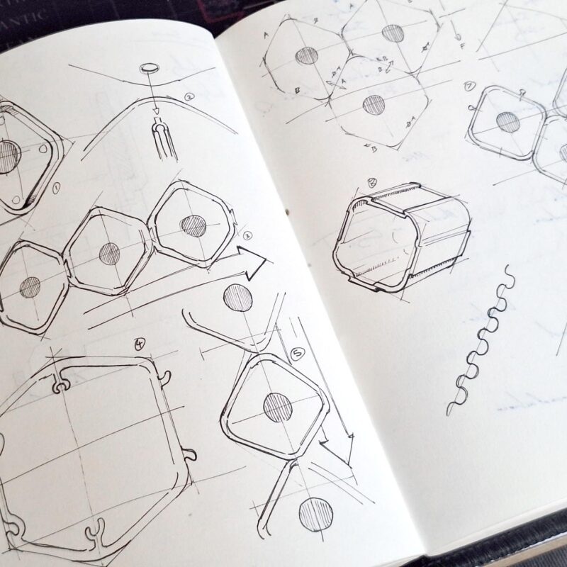 An open sketchbook displays technical line drawings of geometric, hexagonal shapes from various angles, with arrows, diagrams, and notations, suggesting mechanical or structural design concepts.