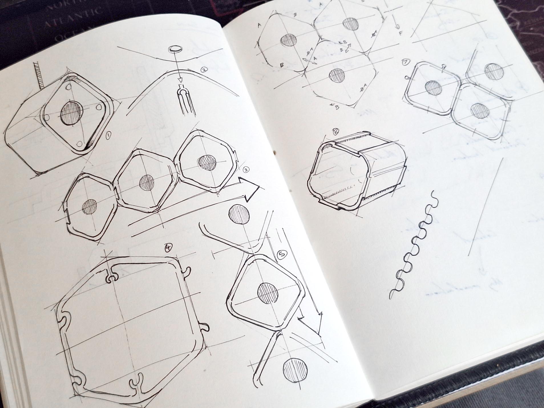 An open sketchbook displays technical line drawings of geometric, hexagonal shapes from various angles, with arrows, diagrams, and notations, suggesting mechanical or structural design concepts.